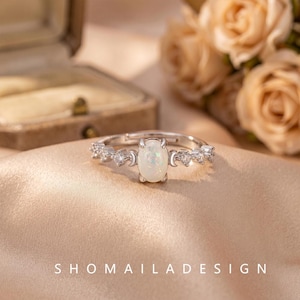 May include: A silver-toned ring featuring a central oval-shaped iridescent opal stone. The band is accented with small clear stones and crescent moon motifs on either side of the centre setting. The text SHOMAILADESIGN is visible at the bottom.