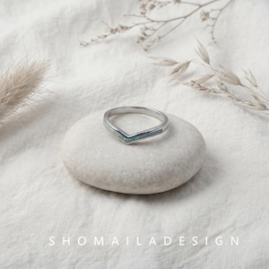 Op de afbeelding: Een zilverkleurige chevronring met een dunne inleg van iriserend blauwgroen opaal. De ring heeft een V-vormig ontwerp en ligt op een gladde, ronde steen. De tekst SHOMAILADESIGN is in het wit onderaan de afbeelding gedrukt.
