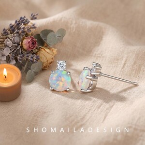 May include: A pair of silver stud earrings featuring round, iridescent opals topped with a small, clear, round-cut gemstone. The earrings are displayed alongside a small lit candle and dried flowers, with the text SHOMAILADESIGN at the bottom.
