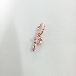 May include: Rose gold-colored charm in the shape of the letter "p". The charm has a small loop at the top for attaching to a bracelet or necklace. A tiny, clear gemstone is set at the top of the letter.