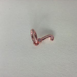 Puede incluir: Charm de color oro rosa con forma de letra "J". El charm presenta una pequeña gema transparente. El diseño incluye un lazo para la fijación y una sección inferior curvada.