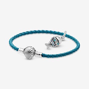 Puede incluir: Una pulsera de cuero trenzado turquesa con un cierre de concha plateada y un dije de pez. El dije de pez es plateado con detalles de esmalte negro, blanco y turquesa. Un accesorio elegante.