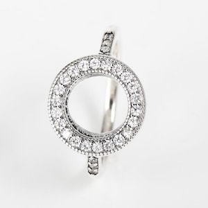 May include: A silver ring with a circular, open centre, surrounded by small, clear gemstones. The band also features small stones. The ring is shown against a white backdrop, highlighting its sparkling details. The ring is a piece of jewellery.