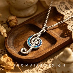 Op de afbeelding: Een zilverkleurige ketting met een vioolsleutel hanger ingelegd met iriserende blauwe en groene steentjes. De hanger zit aan een fijne schakelketting en de tekst SHOMAILDESIGN staat onderaan in de afbeelding gedrukt.