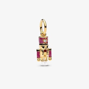 May include: A gold-colored charm in the shape of a toy soldier nutcracker. The nutcracker has a pink glittery hat and sleeves, white boots, and a star-shaped gem. The charm is designed to be hung from a bracelet or necklace.