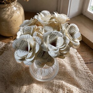 Puede incluir: Un ramo de flores de papel hechas con páginas de libros, dispuestas en un pequeño jarrón de cristal transparente. Las flores son de color crema con texto visible, y el jarrón descansa sobre una tela beige.