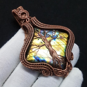 May include: A square labradorite gemstone pendant encased in intricate copper wire wrapping. The wire forms a tree design over the stone, which displays iridescent yellow and blue flashes. The pendant features a decorative wire bail at the top.