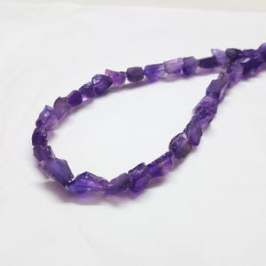 May include: A strand of raw, unpolished amethyst beads in varying shades of deep purple. The beads are irregular in shape and size, featuring a translucent quality that allows light to pass through the natural crystalline structure of the stones.