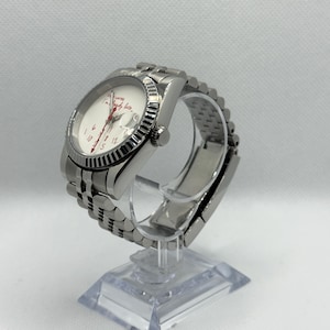 Puede incluir: Un reloj de pulsera plateado con esfera blanca y agujas y números rojos. El reloj tiene una correa plateada y un bisel estriado. La esfera del reloj tiene el texto "I'm ready late" en rojo. El reloj se exhibe sobre un soporte transparente.