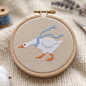 May include: A cross-stitch embroidery hoop featuring a white goose with a blue scarf and orange feet. The goose is set against a beige fabric background within a wooden hoop. Other items include spools of thread and scissors.