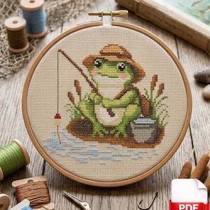 May include: Cross-stitch embroidery of a frog fishing, set in a wooden hoop. The frog is green and wearing a hat and backpack. The scene includes cattails, water, and a small bucket. Spools of thread, scissors, and a PDF icon are in the background.
