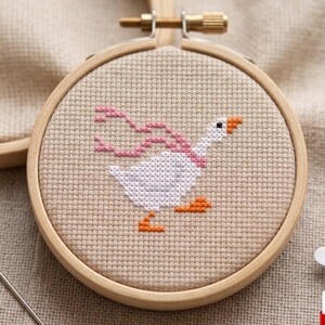 Op de afbeelding: Een borduurring met kruissteek met een witte gans met oranje poten en snavel, met een roze sjaal. De gans is geborduurd op beige stof, gezet in een houten ring. Een naald en draad zijn zichtbaar.