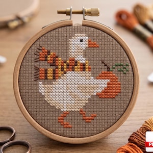 Könnte beinhalten: Ein Kreuzstich-Stickrahmen mit einer weißen Gans, die einen Schal trägt und einen Kürbis hält. Die Gans ist mit weißem, orangefarbenem und braunem Garn auf braunem Stoff gestickt. Der Holzrahmen wird mit einer Messingschraube befestigt.