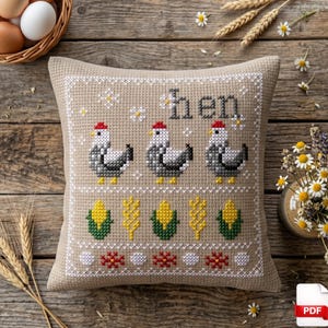 May include: A beige cross-stitched pillow with the word "hen" above three chickens wearing red hats. Below the chickens are corn and wheat designs. The pillow is on a wooden surface with eggs and flowers.