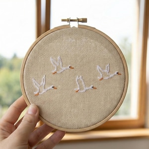 May include: A round, beige cross-stitch embroidery hoop featuring four white geese in flight. The geese have orange beaks and feet. The embroidery is set in a wooden hoop, and the background is a window with natural light.