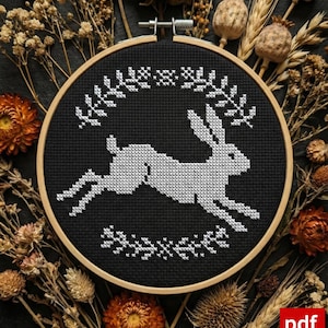 May include: A cross-stitch embroidery hoop featuring a white rabbit leaping across a black background. The design is framed by a wooden hoop and surrounded by dried flowers and seed pods. The rabbit is surrounded by a floral design.