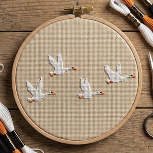 May include: A cross-stitch embroidery hoop featuring four white geese in flight, with orange beaks, against a beige background. Embroidery floss in white, orange, and black, along with small scissors, are arranged around the hoop on a wooden surface.