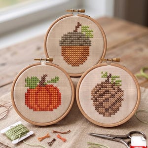 May include: Three cross-stitch embroidery hoops with autumnal designs: an acorn, a pumpkin, and a pinecone. Each design is stitched on beige fabric and set in a wooden hoop. Green and orange threads, plus scissors, are also visible.
