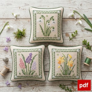 Pode incluir: Três almofadas decorativas quadradas, creme, com desenhos florais bordados em ponto cruz. Cada almofada tem uma borda verde e apresenta diferentes flores: snowdrops, jacintos e narcisos. As almofadas estão dispostas sobre uma superfície de madeira clara com detalhes florais adicionais.