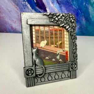 May include: A rectangular silver-toned metal picture frame featuring a three-dimensional cat figure perched on the lower edge and a cluster of sculpted leaves in the upper right corner. The frame holds a print of a cat looking out a window.