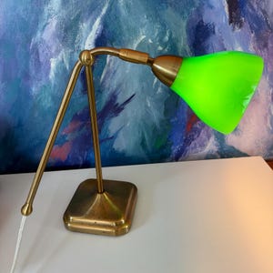 May include: A desk lamp with a brass-toned metal base and an adjustable arm, featuring a vibrant neon green conical glass shade. The shade displays a subtle embossed star pattern on its surface, and the lamp includes a white power cord.