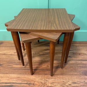 May include: Set of three nesting tables with a mid-century modern design. The tables have a brown wood grain finish and tapered legs. The largest table is approximately 24 inches wide, 16 inches deep, and 20 inches tall.