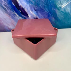 Puede incluir: Una caja de cerámica color rosa empolvado con tapa a juego. La caja tiene forma cuadrada con lados angulados, y la tapa está ligeramente entreabierta, revelando el interior. El fondo presenta una pintura abstracta azul y blanca.