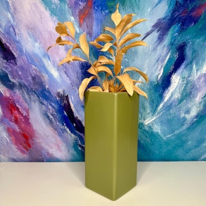 Puede incluir: Un jarrón cuadrado verde oliva alto contiene follaje seco de color marrón dorado. El jarrón se encuentra frente a una pintura abstracta colorida con pinceladas azules, moradas y blancas. El jarrón es una pieza decorativa moderna.