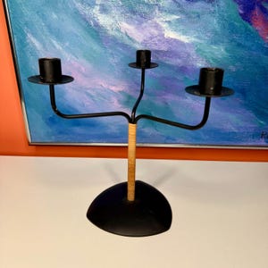 May include: Black metal candelabra with three candle holders. The base is a black, rounded triangle shape. The center stem is wrapped in a light-colored woven material. The background features a colorful abstract painting.