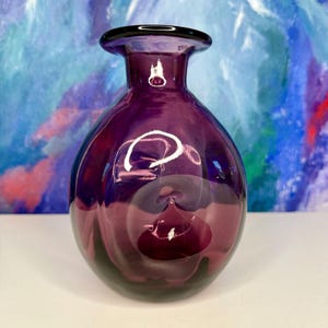 Puede incluir: Un jarrón de cristal morado oscuro con un cuerpo redondeado y un cuello acampanado. El jarrón tiene un borde oscuro y una superficie lisa y reflectante. El fondo presenta una pintura abstracta con tonos azules, morados y rojos.