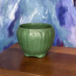 May include: A small, round, sage green ceramic bowl with a fluted, vertical ribbed texture along the exterior. The bowl features a wide, open top and rests on a base with small, integrated feet, showcasing a matte finish and consistent color.