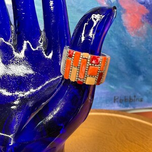 Könnte beinhalten: Ein breiter Ring im Mosaikstil mit einem Muster aus orangefarbenen, roten und cremefarbenen rechteckigen Steinen. Der Ring hat ein silberfarbenes Band und wird auf einer blauen Glashand präsentiert. Das Design des Rings ist geometrisch und auffällig.