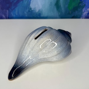 May include: A ceramic seashell-shaped bank in shades of blue and white. The bank has a slot for inserting coins and a glossy finish. The design mimics the natural form of a conch shell, with a dark blue tip.