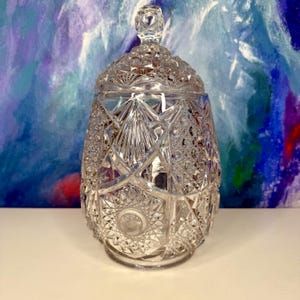 Puede incluir: Un tarro de cristal tallado transparente con tapa. El tarro tiene forma redondeada con intrincados patrones geométricos y un adorno decorativo en la tapa. El cristal refleja la luz. El tarro está sobre una superficie blanca.