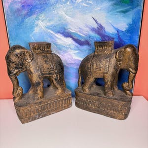 May include: Two antique-style elephant statues with gold and black accents. Each elephant has detailed carvings and a decorative saddle. The statues are displayed on a white surface with a colorful abstract painting in the background.