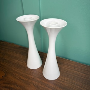 May include: Two white ceramic candle holders with a flared top and a slender, elongated body. The candle holders are placed on a dark wooden surface against a teal-colored wall. The design is minimalist and modern.