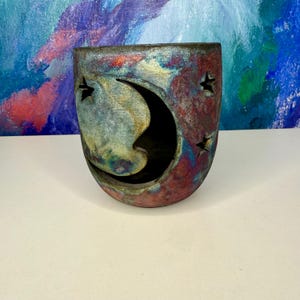 May include: A ceramic candle holder with a celestial design. The holder features a crescent moon cutout and star-shaped cutouts. The color scheme includes shades of red, blue, and gold, creating a unique, artistic appearance.