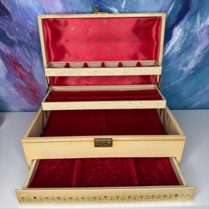 May include: Open vintage jewelry box with multiple compartments and a drawer. The box is cream-colored with a red velvet interior. The lid and drawer are open, revealing the interior storage spaces. The box has a gold-colored clasp and decorative trim.