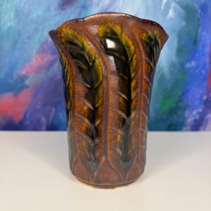 May include: A cylindrical ceramic vase featuring a rich, mottled brown glaze with deep, glossy black and olive green leaf-like vertical patterns carved into the surface. The rim has a scalloped, wavy edge, and the body has a textured finish.