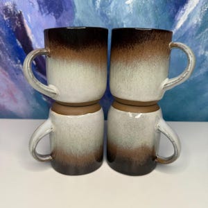 May include: Four ceramic mugs stacked in pairs. Each mug has a rounded body and a curved handle. The mugs feature a gradient color scheme, transitioning from dark brown at the base to white in the middle, and dark brown at the top.