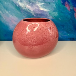 Könnte beinhalten: Eine runde, rosa Keramikvase mit gesprenkelter Textur. Die Vase hat eine weite Öffnung und eine glänzende Oberfläche. Der Hintergrund zeigt ein farbenfrohes abstraktes Gemälde mit Blau- und Grüntönen. Diese dekorative Vase eignet sich für die Inneneinrichtung.