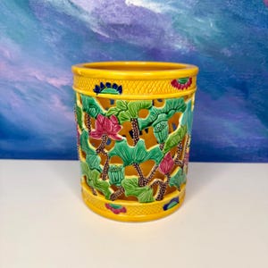 May include: A yellow ceramic vase with a detailed lotus flower design. The vase has a cylindrical shape with cut-out patterns of green leaves, pink flowers, and brown stems. The top and bottom rims are yellow with blue and pink accents.