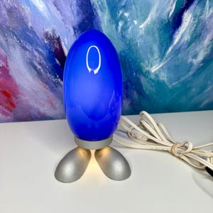 May include: A table lamp featuring a vibrant blue, egg-shaped glass shade supported by two silver-colored, foot-shaped metal bases. The lamp includes a white electrical cord with an inline switch, providing a steady light source from the base.