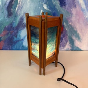 May include: A wooden table lamp with a rectangular shade. The lamp has a warm, natural wood finish and a colorful, abstract design on the shade. The lamp is plugged in with a black cord.