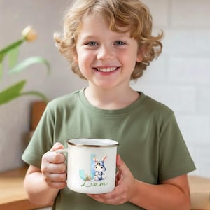 Könnte beinhalten: Weißer Emaillebecher mit silbernem Rand. Der Becher zeigt den blauen Buchstaben "J" und eine Aquarellillustration eines Hasen mit Hut. Der Name "Liam" steht darunter in Schreibschrift. Der Becher wird von einem Kind gehalten.