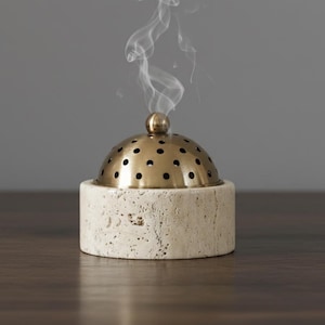 Puede incluir: Un quemador de incienso de piedra beige con una cúpula de latón perforada y un pequeño pomo de latón. Volutas de humo blanco se elevan del quemador. El quemador está sobre una superficie de madera oscura.