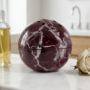 May include: A spherical, burgundy marble container with white veining. The container has a lid and several small holes on top. A clear glass bottle with yellow liquid is to the left, and a white rose is to the right.