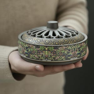 May include: A gray ceramic incense burner with a decorative lid featuring a series of arched openings. The body is adorned with a colorful floral and scroll pattern in green, pink, and gold. The burner is held in a person's hands.