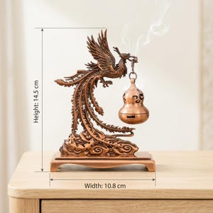 Op de afbeelding: Een bronskleurige wierookbrander in de vorm van een feniks, 14,5 cm hoog en 10,8 cm breed, met een kalebasvormige wierookhouder. De feniks staat op een decoratieve basis, met rook die opstijgt uit de wierookhouder. Het item staat op een houten oppervlak.