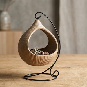 May include: A beige ceramic hanging planter with a textured finish, supported by a black metal stand. The teardrop-shaped planter contains dried plants, suitable for indoor decoration. The stand features a circular base and a curved arm.